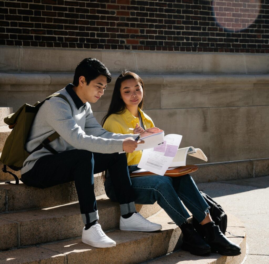 A young man and woman reviewing papers while sitting on outdoor steps, enjoying a sunny day.